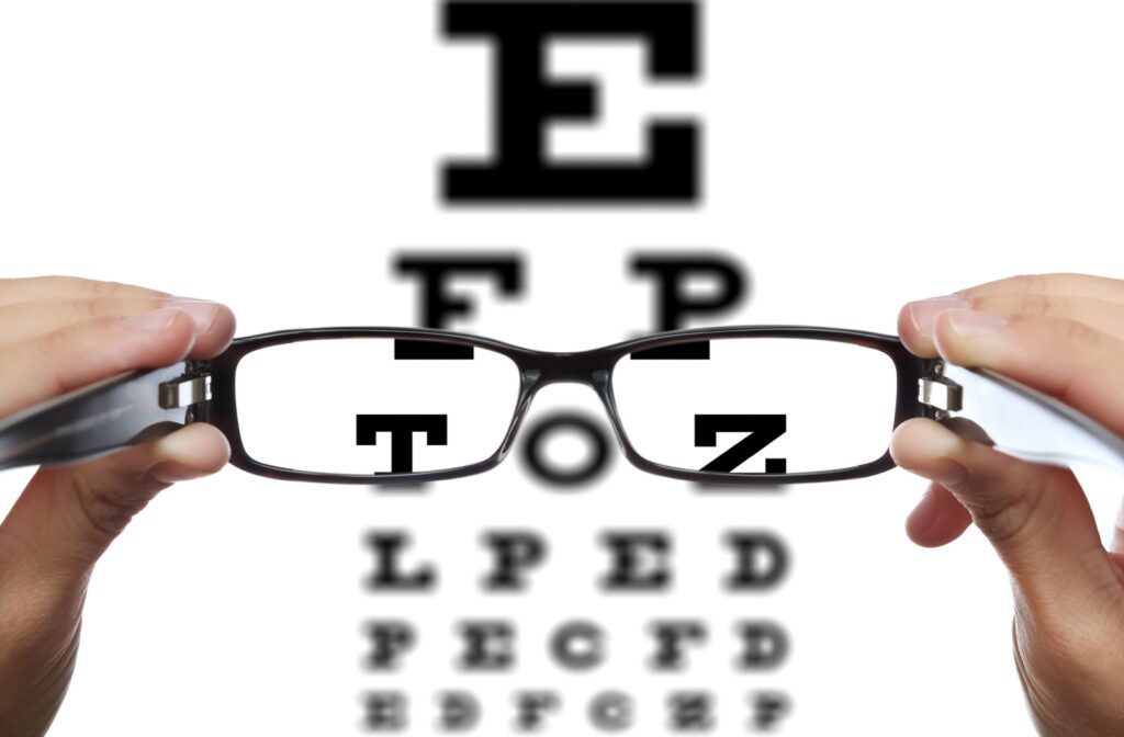 In front of a blurry visual acuity test, a pair of hands holds up eyeglasses that sharpen some of the letters.