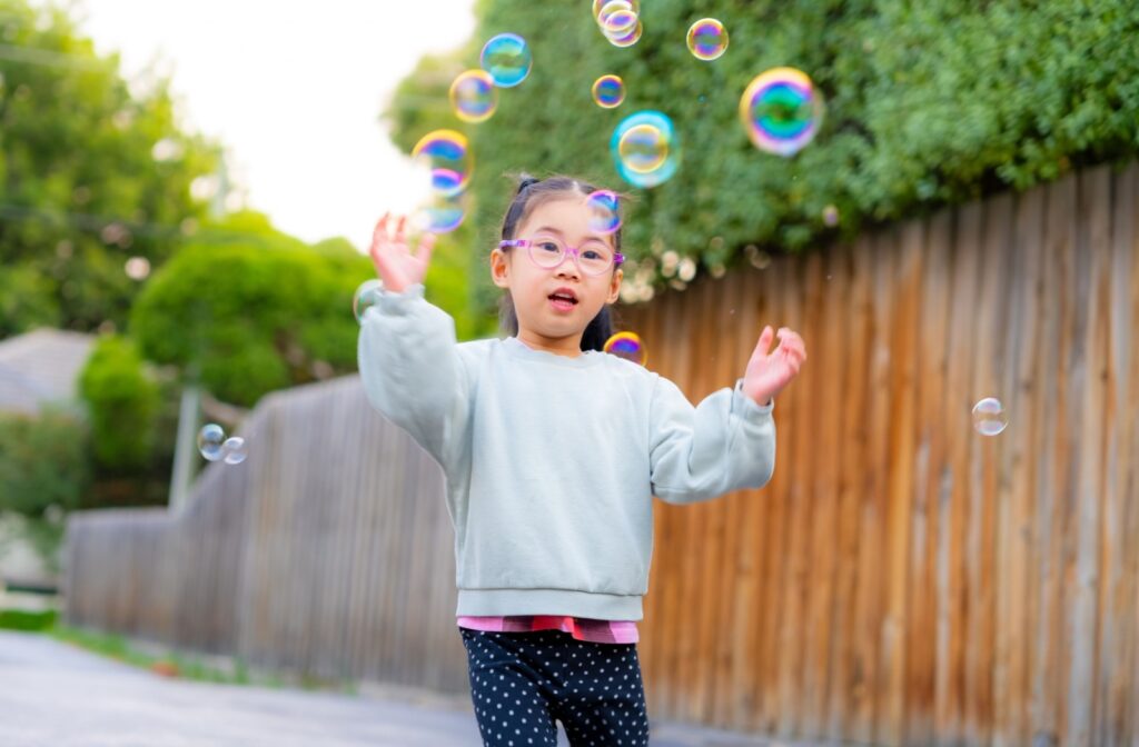 A young child in pink glasses plays outside with iridescent bubbles as part of their myopia management plan.