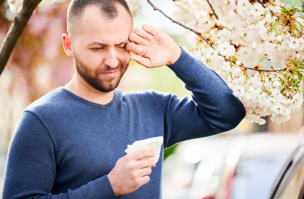 Man rubbing his itchy eyes while standing near blooming trees during allergy season.